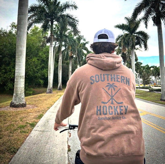 Brown Southern Hockey hoodie lifestyle photo from back side on sidewalk with palm trees on both sides of the person on a bike