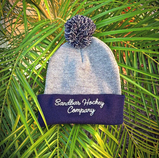 Gray and navy hockey beanie lifestyle image with pom pom and Sandbar Hockey Company cursive script logo