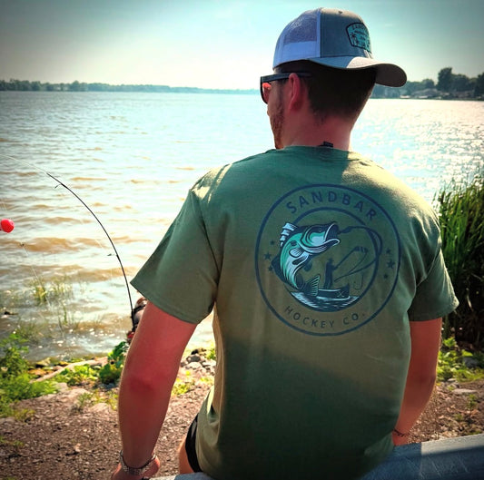Military green hockey t-shirt lifestyle photo from back side by the water featuring the bass fishing graphic