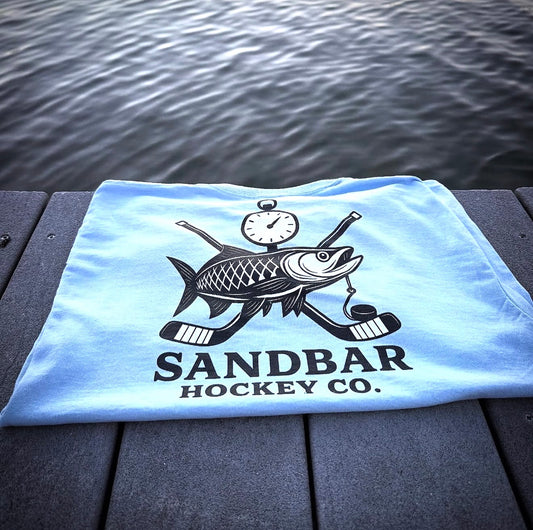 Sandbar Hockey Fishing Edition folded light blue t-shirt with fish, crossed hockey sticks, and stopwatch graphic