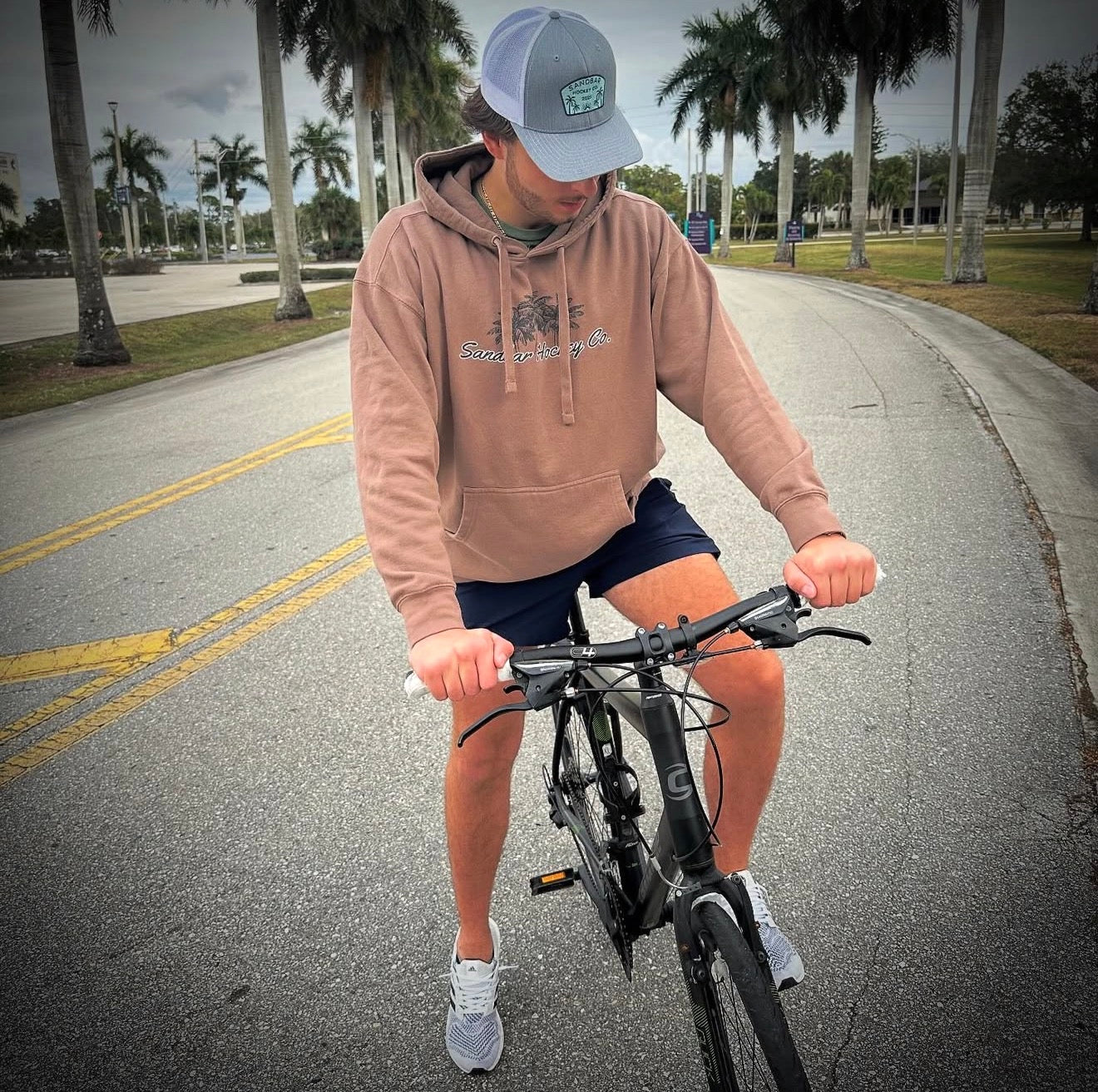 Brown Southern Hockey hoodie lifestyle photo from front side on bicycle with palm trees in the backround