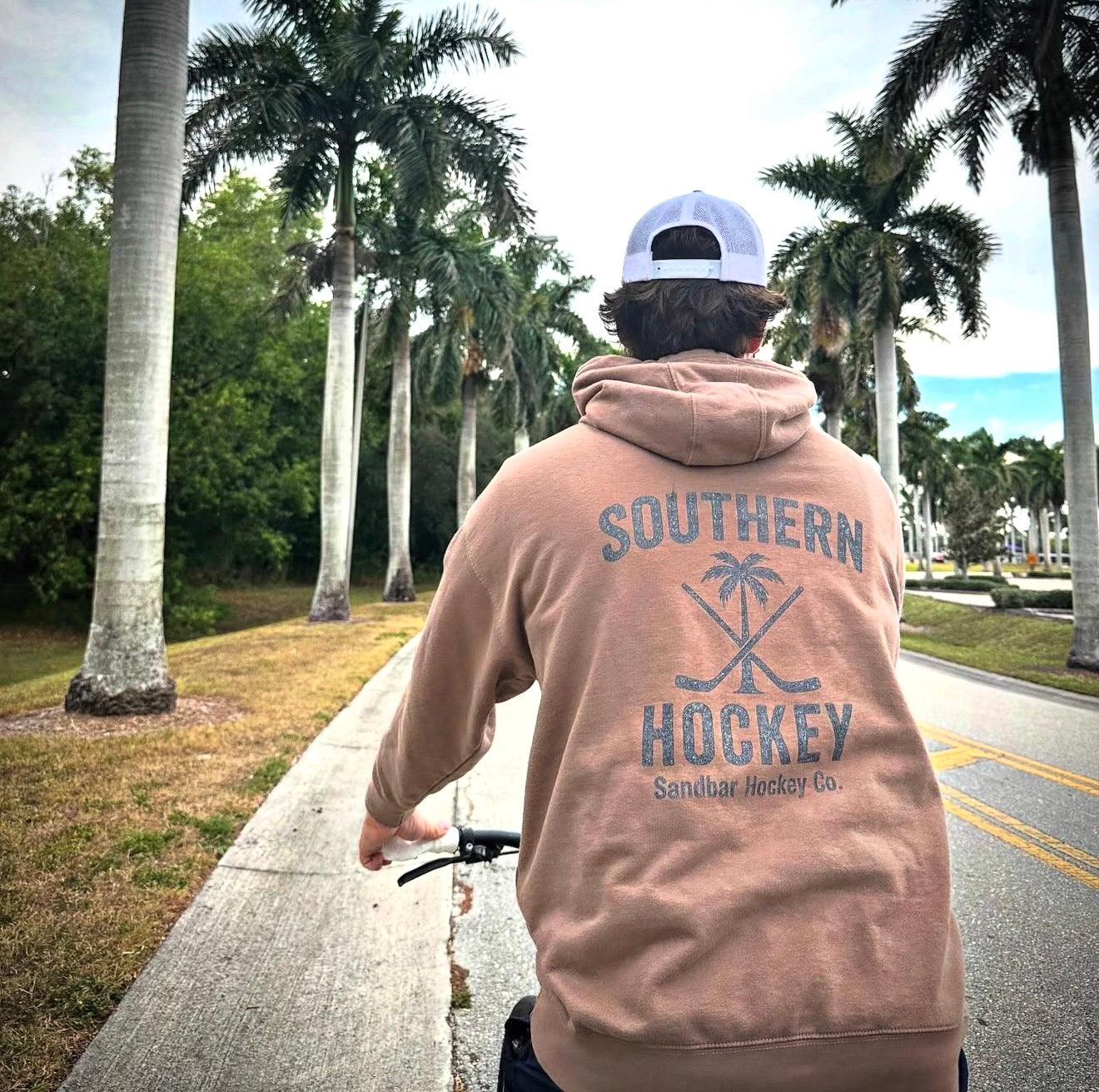 Brown Southern Hockey hoodie lifestyle photo from back side on sidewalk with palm trees on both sides of the person on a bike