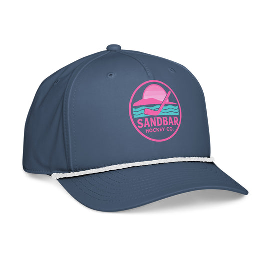 Blue women’s hockey rope hat left front angle view with pink Sandbar Hockey Co. sunset logo, white rope detail, and curved brim.
