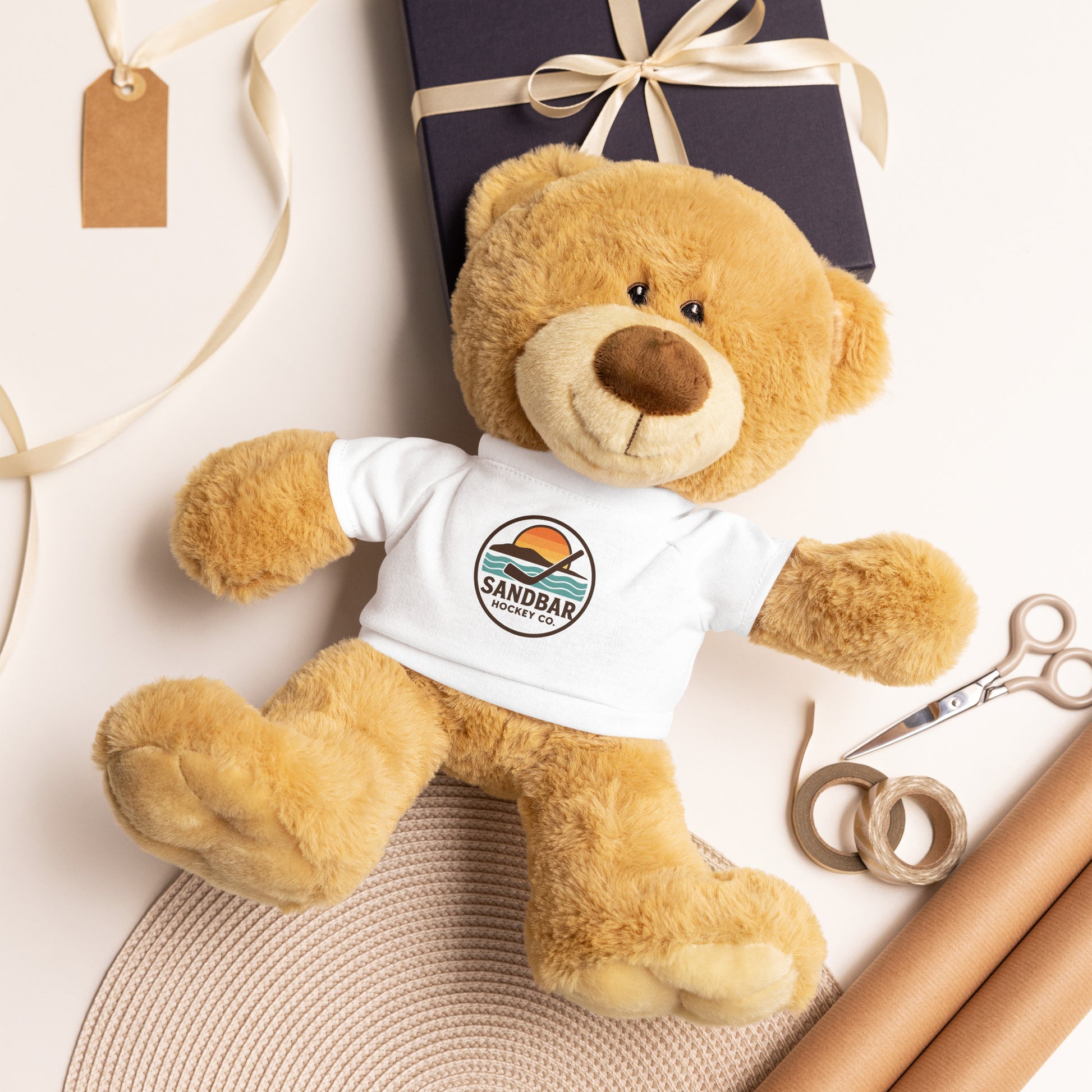 Tan teddy bear wearing a white Sandbar Hockey Co. t-shirt with sunset logo, styled on a gift wrapping background with ribbon, scissors, and wrapping paper.